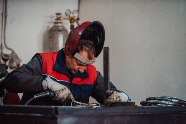 Profesional welder in protective uniform and mask welding metal pipe on the industrial table with other workers behind in the industrial workshop