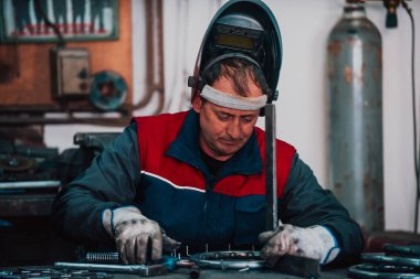 Close up welder in protective uniform and mask welding metal pipe on the industrial table with other workers behind in the industrial workshop