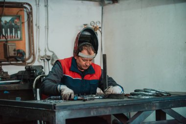 Profesional welder in protective uniform and mask welding metal pipe on the industrial table with other workers behind in the industrial workshop