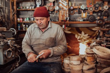 A senior man sitting in the workshop and processing wooden utensils in the old manual way. 