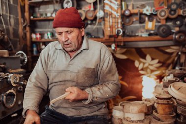 A senior man sitting in the workshop and processing wooden utensils in the old manual way. 