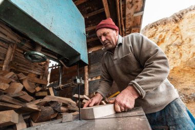 A senior man processing wood on a machine in an outdoor workshop. 