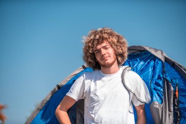A man camping with a tent. He is surrounded by a beautiful mountain landscape, and the sun shines on his face, foreshadowing a beautiful and sunny day. The man looks satisfied and fulfilled