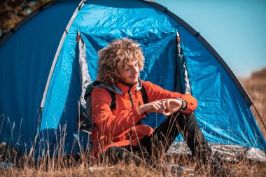 A man camping with a tent. He is surrounded by a beautiful mountain landscape, and the sun shines on his face, foreshadowing a beautiful and sunny day. The man looks satisfied and fulfilled