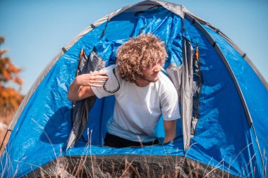 A man with a tent enjoys a sunny day whileusing a smartphone and camping in the mountains.