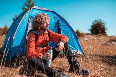 A man camping with a tent. He is surrounded by a beautiful mountain landscape, and the sun shines on his face, foreshadowing a beautiful and sunny day. The man looks satisfied and fulfilled