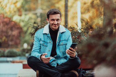 A gappy student sitting in a park using a smartphone and wireless headphones. 