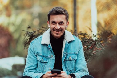 A gappy student sitting in a park using a smartphone and wireless headphones. 