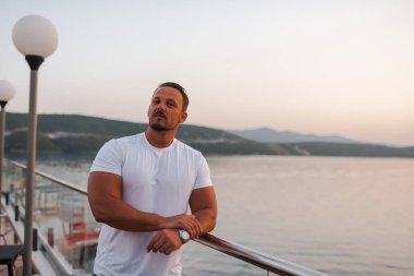 Kumlu bir sahil yakınındaki bir otelde tatil yapan fit ve çekici bir adam. O şık bir şapka takıyor ve sıcak güneşin ve kristal berrak denizin tadını çıkarıyor. Manzara huzurlu ve huzurlu, bir duyguyu yansıtıyor.