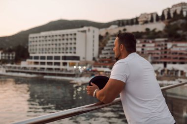 Kumlu bir sahil yakınındaki bir otelde tatil yapan fit ve çekici bir adam. O şık bir şapka takıyor ve sıcak güneşin ve kristal berrak denizin tadını çıkarıyor. Manzara huzurlu ve huzurlu, bir duyguyu yansıtıyor.