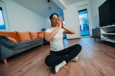 Genç bir kadın yoga yapıyor ve kafasında kulaklıklarla modern bir dairede dinleniyor..