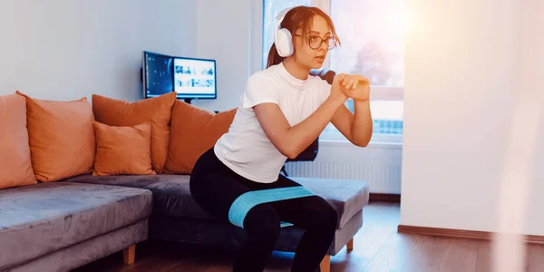 A young woman doing squats in a modern apartment, wearing headphones on her head, indicating shes listening to music or an audio workout guide. Her determined expression and active pose showcase her
