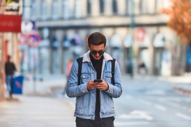 Şehirde gezen bir öğrenci bir sırt çantası taşıyor ve akıllı telefon kullanırken güneş gözlüğü takıyor, modern şehir yaşam tarzını ve iletişim ve güvenilirliği temsil ediyor.