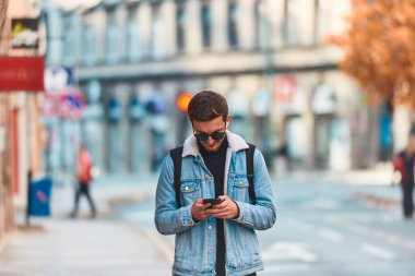 Şehirde gezen bir öğrenci bir sırt çantası taşıyor ve akıllı telefon kullanırken güneş gözlüğü takıyor, modern şehir yaşam tarzını ve iletişim ve güvenilirliği temsil ediyor.