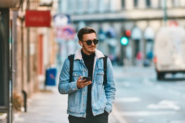 Şehirde gezen bir öğrenci bir sırt çantası taşıyor ve akıllı telefon kullanırken güneş gözlüğü takıyor, modern şehir yaşam tarzını ve iletişim ve güvenilirliği temsil ediyor.