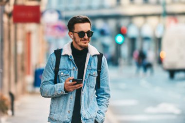 Şehirde gezen bir öğrenci bir sırt çantası taşıyor ve akıllı telefon kullanırken güneş gözlüğü takıyor, modern şehir yaşam tarzını ve iletişim ve güvenilirliği temsil ediyor.