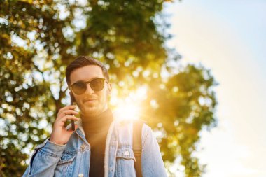 Şehirde gezen bir öğrenci bir sırt çantası taşıyor ve akıllı telefon kullanırken güneş gözlüğü takıyor, modern şehir yaşam tarzını ve iletişim ve güvenilirliği temsil ediyor.