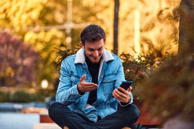 Şehir merkezindeki bankta oturan bir adam telefon etmek için akıllı telefonu ve kablosuz kulaklığını kullanıyor, modern iletişimde teknolojinin kolaylığını ve aynı anda birden fazla iş yapabilme yeteneğini sergiliyor.