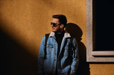 Bir adam turuncu bir duvarın önünde duruyor, güneş gözlüğü, sırt çantası ve kablosuz kulaklık takıyor, modern bir şehir tarzı yayıyor, bağımsızlık ve kendine güven hissi veriyor.