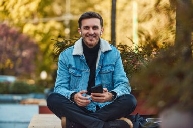 Şehir merkezindeki bankta oturan bir adam telefon etmek için akıllı telefonu ve kablosuz kulaklığını kullanıyor, modern iletişimde teknolojinin kolaylığını ve aynı anda birden fazla iş yapabilme yeteneğini sergiliyor.