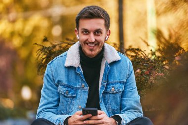 Şehir merkezindeki bankta oturan bir adam telefon etmek için akıllı telefonu ve kablosuz kulaklığını kullanıyor, modern iletişimde teknolojinin kolaylığını ve aynı anda birden fazla iş yapabilme yeteneğini sergiliyor.