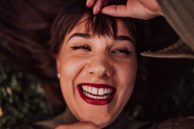 Genç bir kadın çimlerin üzerinde uzanıyor, güzel yüzünü sakin bir ifadeyle gösteriyor. Arka plandaki yemyeşil yeşillik sahnenin huzurlu atmosferini artırıyor ve rahatlatıyor.