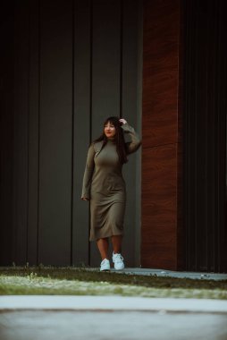 Kendine güvenen ve tarz sahibi bir kadın zeytin yeşili bir elbiseyle modern bir binanın önünden geçiyor. Işıl ışıl bir güzellik, güç ve kendini beğenmişlik.