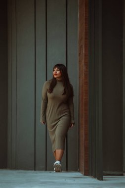 Kendine güvenen ve tarz sahibi bir kadın zeytin yeşili bir elbiseyle modern bir binanın önünden geçiyor. Işıl ışıl bir güzellik, güç ve kendini beğenmişlik.