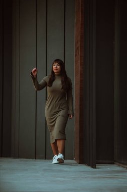 Kendine güvenen ve tarz sahibi bir kadın zeytin yeşili bir elbiseyle modern bir binanın önünden geçiyor. Işıl ışıl bir güzellik, güç ve kendini beğenmişlik.