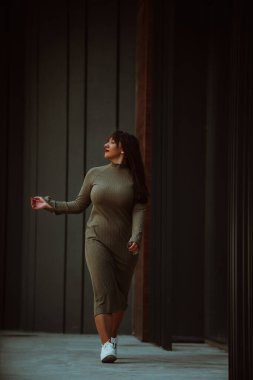 Kendine güvenen ve tarz sahibi bir kadın zeytin yeşili bir elbiseyle modern bir binanın önünden geçiyor. Işıl ışıl bir güzellik, güç ve kendini beğenmişlik.