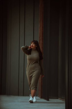 Kendine güvenen ve tarz sahibi bir kadın zeytin yeşili bir elbiseyle modern bir binanın önünden geçiyor. Işıl ışıl bir güzellik, güç ve kendini beğenmişlik.
