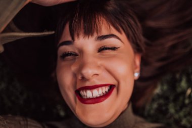 Genç bir kadın çimlerin üzerinde uzanıyor, güzel yüzünü sakin bir ifadeyle gösteriyor. Arka plandaki yemyeşil yeşillik sahnenin huzurlu atmosferini artırıyor ve rahatlatıyor.