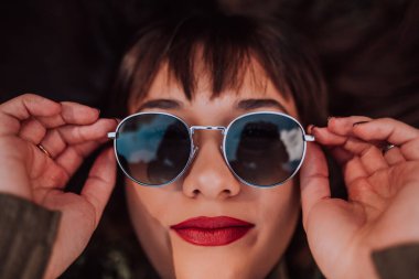 Güneş gözlüklü genç bir kadın çimlerin üzerinde uzanıyor, güzel yüzünü sakin bir ifadeyle gösteriyor.