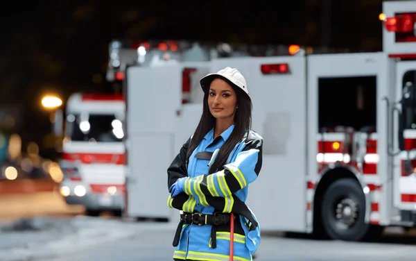 A portrait of a woman in a firefighter uniform with crossed arms, background of a nighttime street where a fire truck is seen after an intervention