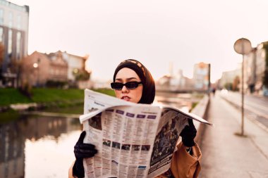 Eski ve modern Fransız kıyafetlerinin birleşimi ile süslenmiş tesettürlü bir kadın, şehrin hareketli sokaklarına dalmış, bir gazete okumaya dalmış.. 