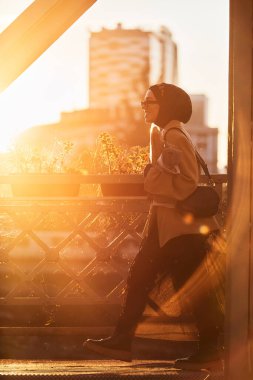 Şık güneş gözlüklü tesettürlü ve zarif Fransız kıyafetli bir kadın, gün batımında şehirde yürüyor, bir buket ekmek ve gazete taşıyor, kültürel bir cazibe ve dinginlik hissi yayıyor.. 