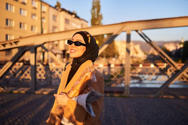 stock image A hijab woman in stylish sunglasses and an elegant French outfit, walking through the city at sunset, carrying a bouquet, bread, and newspaper, radiating a sense of cultural charm and serenity. 