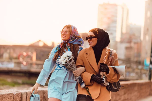 ヒジャーブと現代的で伝統的なドレスを着たカップルの女性と 青いドレスとスカーフを着たカップルは 日没時に街を歩いています 花束とパンを持っているのに対し — ストック写真