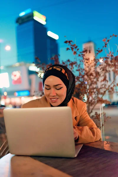 stock image In the enchanting ambiance of a nocturnal cityscape, a hijab-clad girl engrossed in her laptop creates a mesmerizing image, embodying the fusion of technology, empowerment, and the vibrant urban