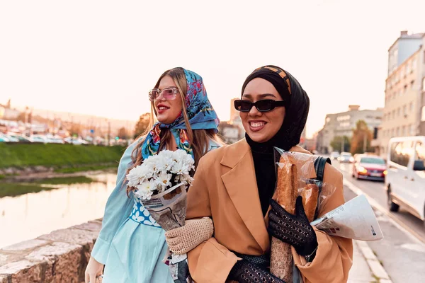 stock image Couple woman one wearing a hijab and a modern yet traditional dress, and the other in a blue dress and scarf, walking together through the city at sunset. One carries a bouquet and bread, while the