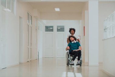 Afro-Amerikan hastane teknisyeni hastane koridorlarında şefkatle geziniyor, tekerlekli sandalyeye mahkum iş arkadaşını itiyor, birlik, destek ve kapsayıcılığı simgeliyor.. 