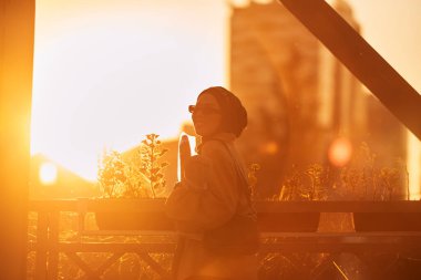 Şık güneş gözlüklü tesettürlü ve zarif Fransız kıyafetli bir kadın, gün batımında şehirde yürüyor, bir buket ekmek ve gazete taşıyor, kültürel bir cazibe ve dinginlik hissi yayıyor.. 