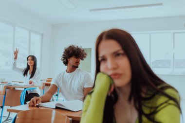 Modern bir sınıfta bir grup farklı öğrencinin önünde oturan yorgun görünüşlü bir öğrenci, akademik yolculuklarında yorgunluk, öğrenme ve işbirliği karışımını yakalıyor..