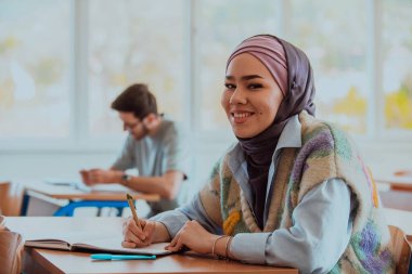 Modern bir sınıfta oturan Müslüman bir tesettür öğrencisi, sınıfı dikkatle takip ederek, bilgi arayan ve eğitime dahil olmayı teşvik eden bir ruhu somutlaştırıyor.. 