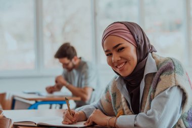 Modern bir sınıfta oturan Müslüman bir tesettür öğrencisi, sınıfı dikkatle takip ederek, bilgi arayan ve eğitime dahil olmayı teşvik eden bir ruhu somutlaştırıyor.. 