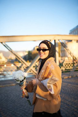 Şık güneş gözlüklü tesettürlü ve zarif Fransız kıyafetli bir kadın, gün batımında şehirde yürüyor, bir buket ekmek ve gazete taşıyor, kültürel bir cazibe ve dinginlik hissi yayıyor.. 