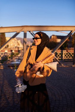 Şık güneş gözlüklü tesettürlü ve zarif Fransız kıyafetli bir kadın, gün batımında şehirde yürüyor, bir buket ekmek ve gazete taşıyor, kültürel bir cazibe ve dinginlik hissi yayıyor.. 