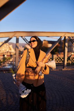 Şık güneş gözlüklü tesettürlü ve zarif Fransız kıyafetli bir kadın, gün batımında şehirde yürüyor, bir buket ekmek ve gazete taşıyor, kültürel bir cazibe ve dinginlik hissi yayıyor.. 