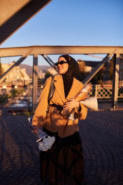 Şık güneş gözlüklü tesettürlü ve zarif Fransız kıyafetli bir kadın, gün batımında şehirde yürüyor, bir buket ekmek ve gazete taşıyor, kültürel bir cazibe ve dinginlik hissi yayıyor.. 