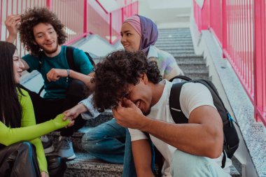 Bir grup öğrenci eğitim molası sırasında modern bir okul koridorunun basamaklarında oturuyor, kahkahalar, espriler ve bilgiler paylaşıyor, birlik ve dostluk duygusunu somutlaştırıyorlar.